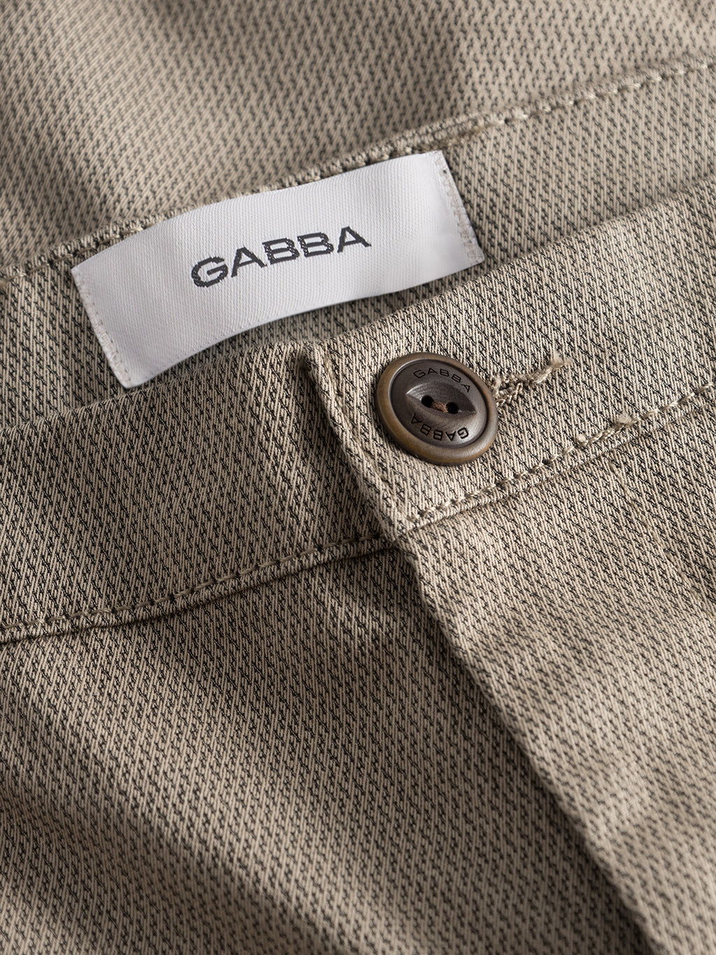Close-up of Pisa K3280 Dale Pants - Humus by GABBA, showing beige textured high-stretch fabric, a white GABBA label, and a brown branded button above the pocket.