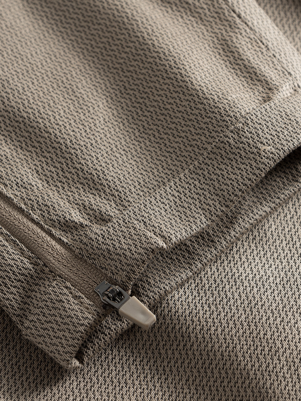 Close-up of textured beige high-stretch fabric with a visible zipper and seam, from the GABBA Pisa K3280 Dale Pants in Humus. The material has a subtle, small-scale repeating pattern for a modern tapered fit.