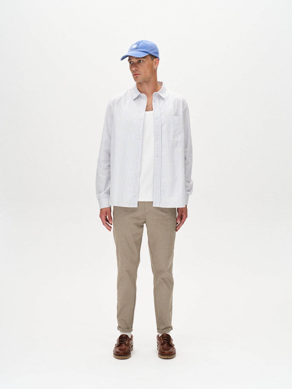 A man stands against a white background wearing the GABBA Pisa K3280 Dale Pants in Humus, paired with a light blue baseball cap, pinstriped button-up over a white T-shirt, and brown boat shoes, looking to his left.