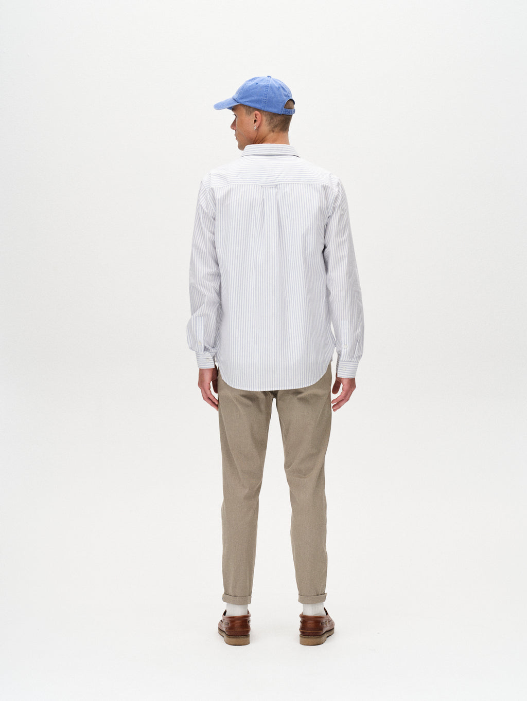 A man stands facing away, wearing a light blue cap, a white striped button-down shirt, GABBA Pisa K3280 Dale Pants in Humus, and brown shoes, against a plain white background.