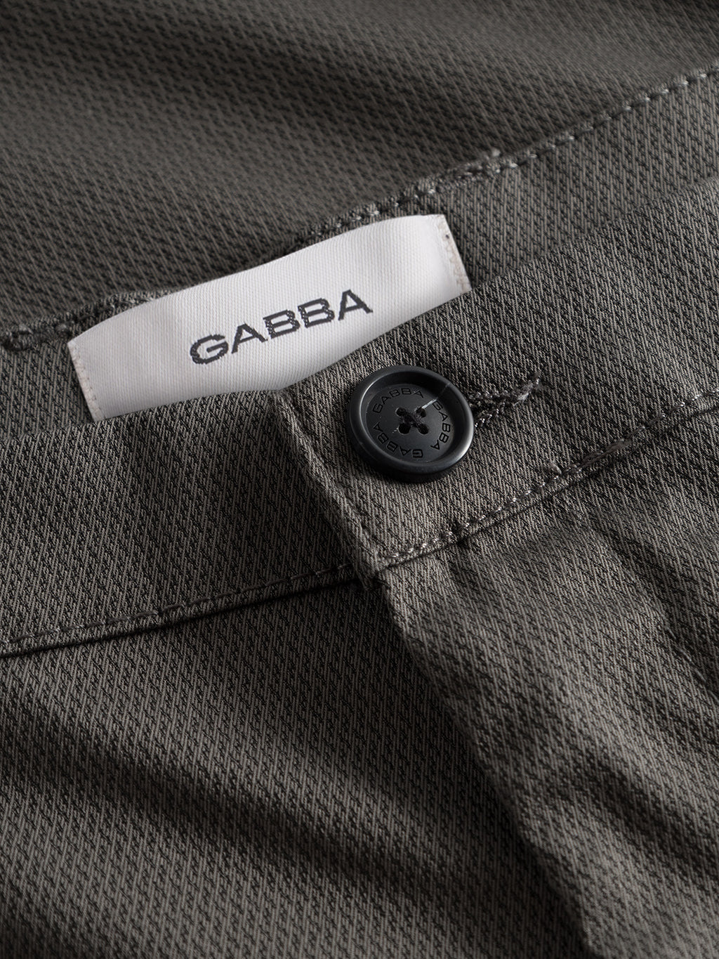 Close-up of GABBAs Pisa K3280 Dale Pants in grey, featuring textured high-stretch cotton-blend fabric, a black button, and a white inner waistband label with GABBA branding.