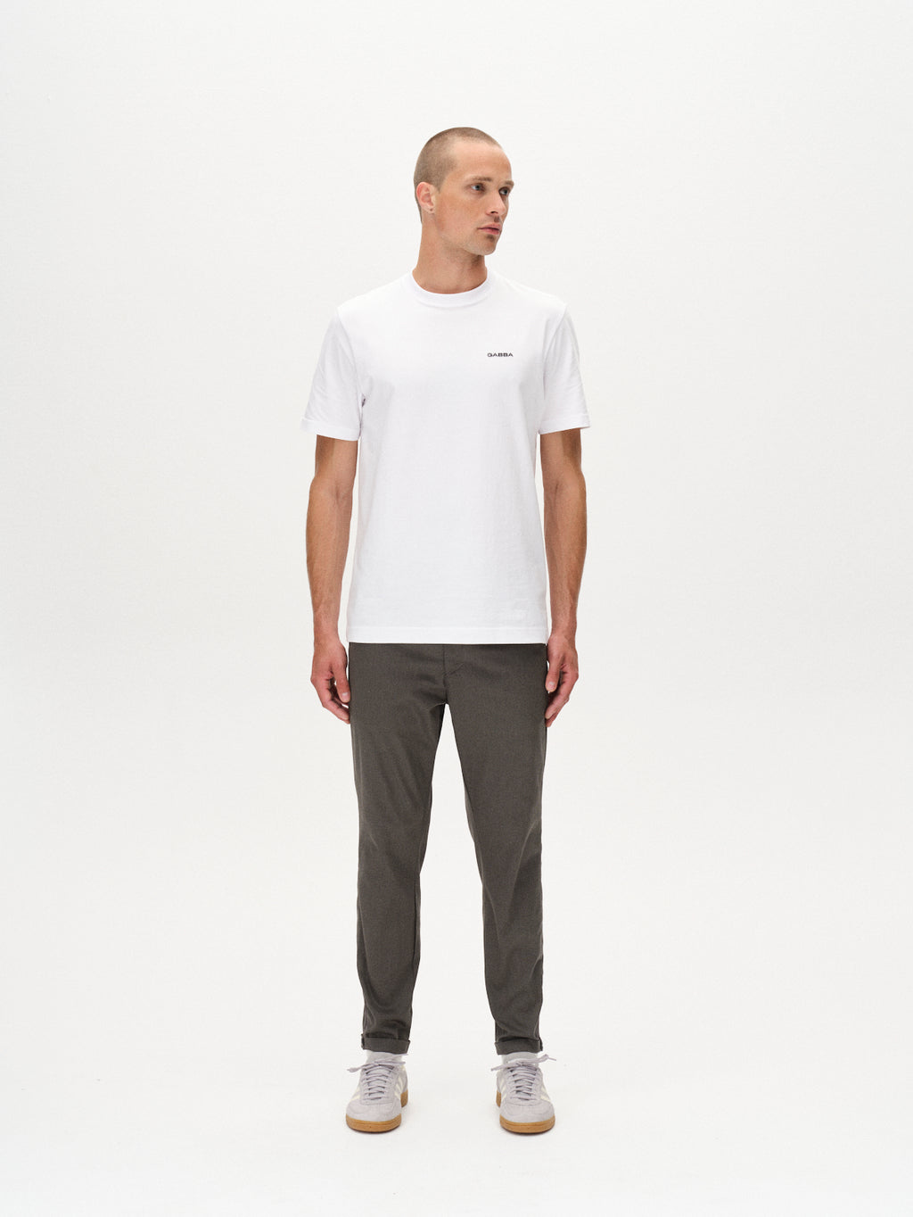 A man with short hair stands against a plain white background, wearing a white t-shirt, light gray sneakers, and GABBA Pisa K3280 Dale Pants in grey made from high-stretch cotton-blend fabric. He looks slightly left with arms relaxed at his sides.
