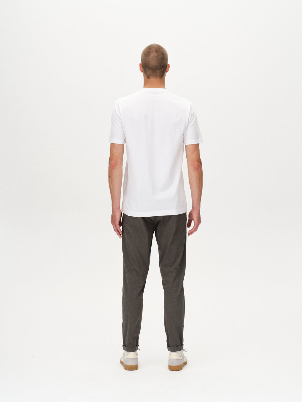 A man faces away from the camera against a white background, wearing a plain white t-shirt, GABBA Pisa K3280 Dale Pants in grey with zippers, and white high-stretch cotton-blend sneakers.