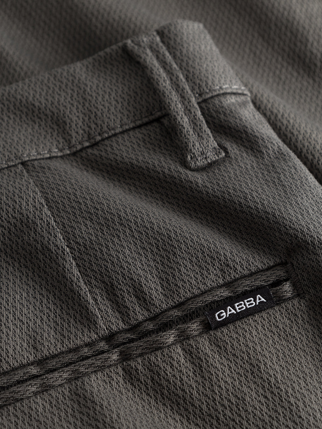Close-up of the GABBA Pisa K3280 Dale Pants in grey, showcasing a tapered fit with belt loops and pockets. A small black label with white GABBA text is next to the pocket seam. Crafted from high-stretch cotton-blend fabric, these modern city pants feature a subtle woven pattern.