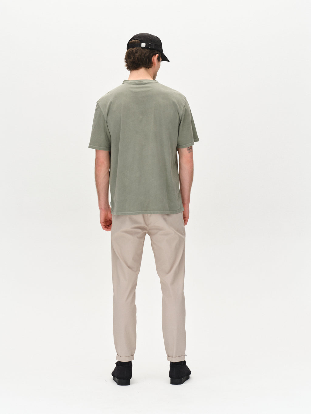 A person stands with their back to the camera, wearing a green T-shirt, Gabbas Pisa K3280 Dale Pants in Grey Paloma, black shoes, and a black cap against a plain light background.