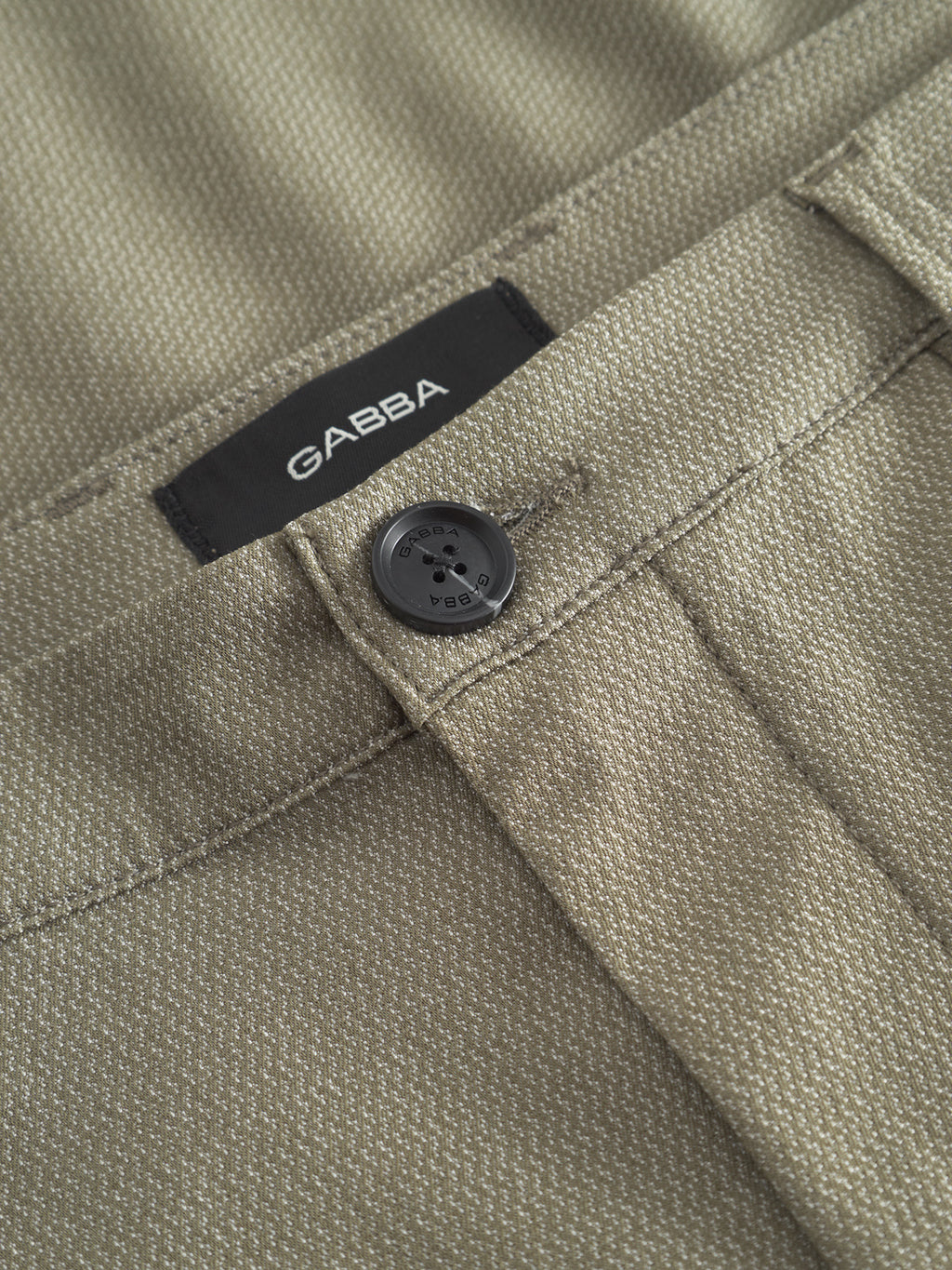 Close-up of olive green Pisa K3280 Dale Pants by Gabba. Made from high-stretch cotton-blend fabric, they feature a black button and label with Gabba in white. The textured weave and prominent waistband stitching offer a sleek, modern city pant look.
