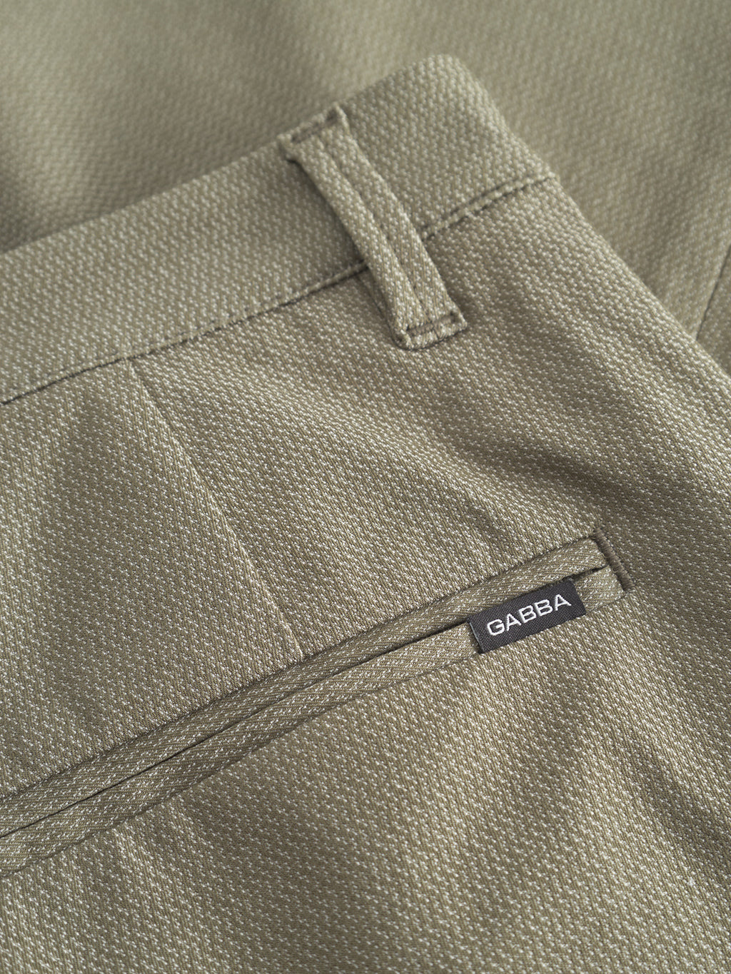 A close-up of the Pisa K3280 Dale Pants in olive green displays a back pocket featuring a classic black and white Gabba label. Made from high-stretch cotton-blend material, these modern city pants include a subtle woven pattern, belt loops, and sleek Gabba Pisa zippers.