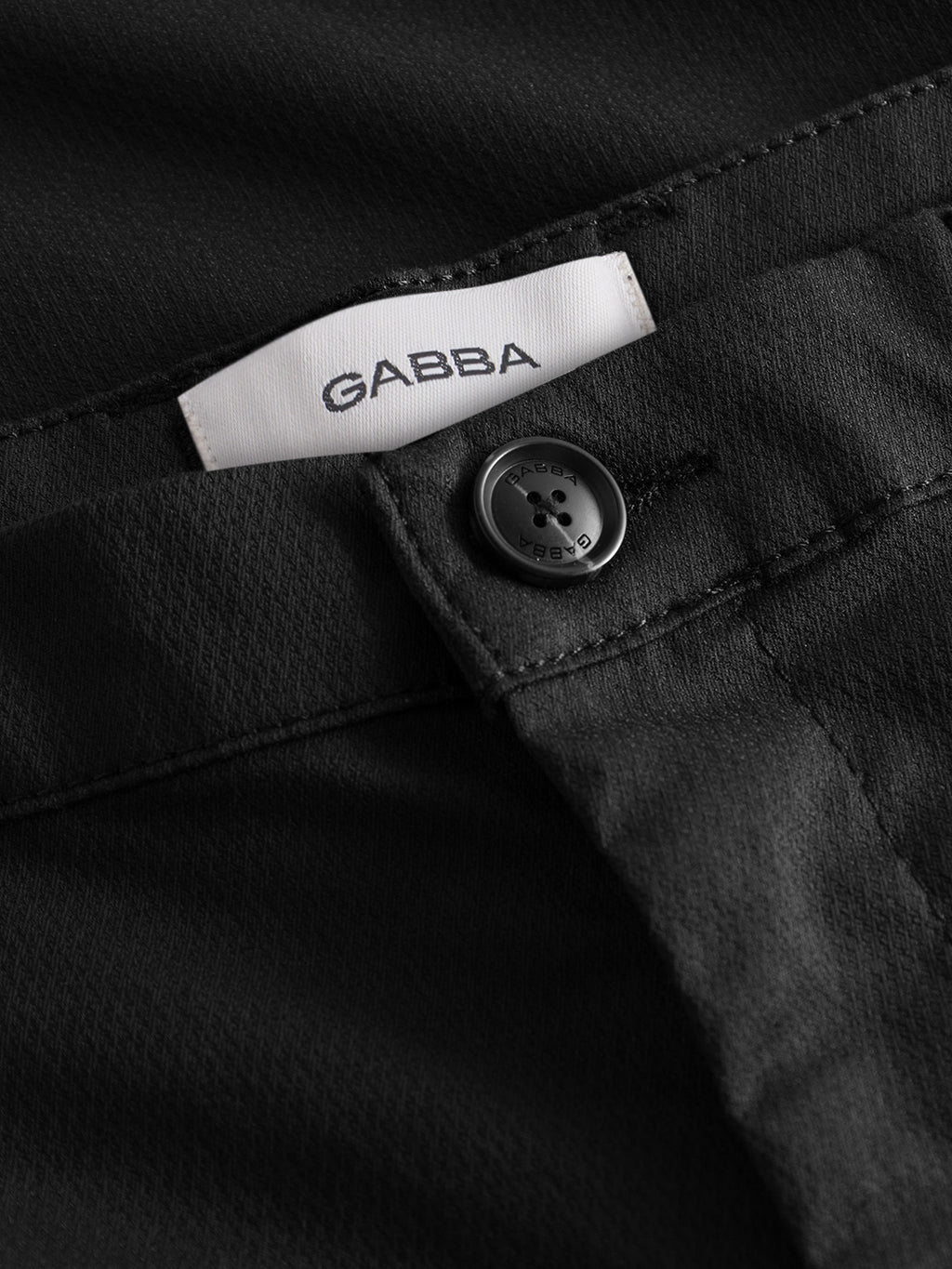 Close-up of Gabba Pisa K3280 Dale Pants in black, featuring a black button, white Gabba label, and subtle pinstripes on high-stretch cotton-blend fabric with visible texture and stitching details.