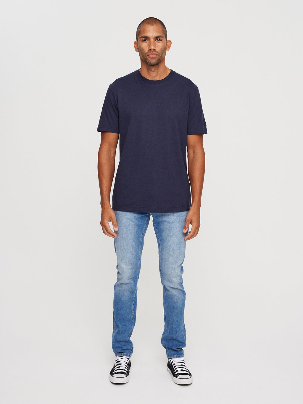 A person with short hair faces the camera in a neutral expression, against a plain backdrop, wearing Gabbas Dune SS GOTS Tee in Total Eclipse made from organic cotton, paired with light blue jeans and black and white sneakers.