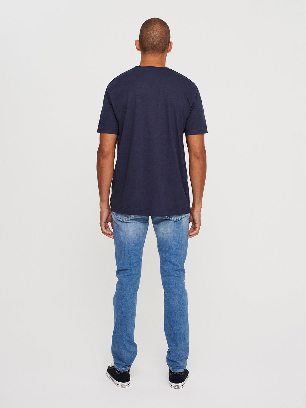 A person is seen from the back wearing a Gabba Dune SS GOTS Tee in Total Eclipse navy blue and light blue jeans. The plain, neutral background highlights their casual, organic cotton outfit.