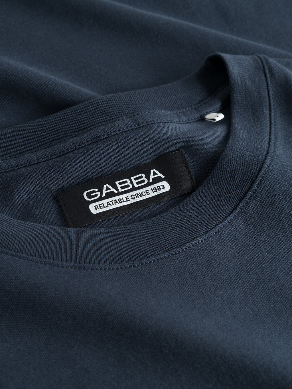 Close-up of a folded navy blue Dune SS GOTS Tee - Total Eclipse, featuring a black Gabba label near the collar reading Relatable Since 1983. Made from organic cotton, the visible fabric texture highlights the quality of this basic t-shirt.