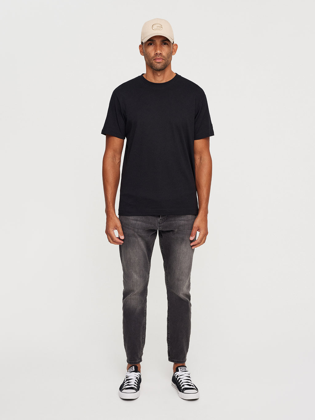 Against a plain background, a person faces forward wearing a Gabba beige cap, Dune SS GOTS Tee in black, gray jeans, and black sneakers with white soles. Their arms rest at their sides.