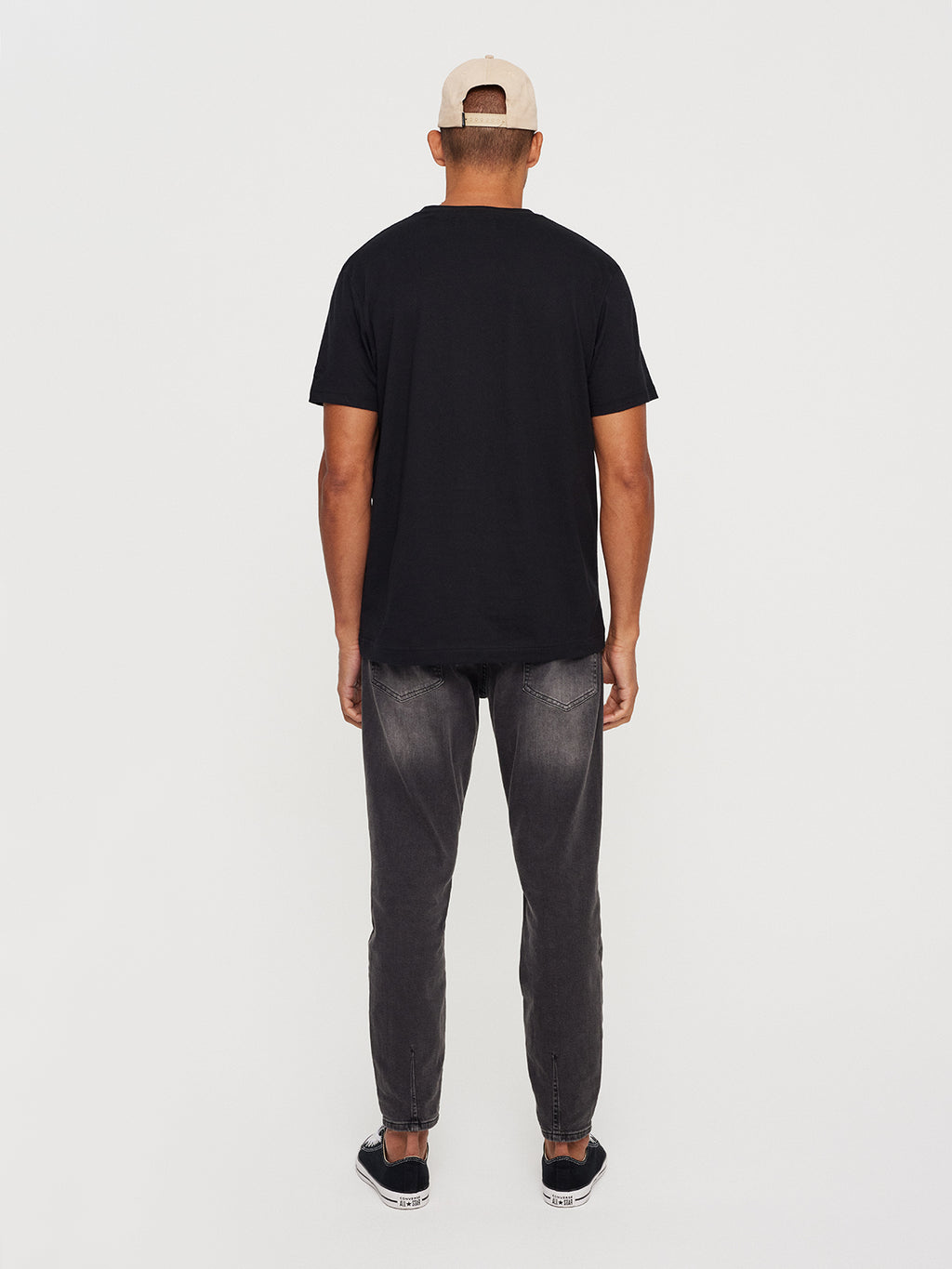 A person stands facing away, wearing a Gabba beige cap, Dune SS GOTS Tee in classic fit black made of organic cotton, faded black jeans, and black sneakers. The background is plain white.