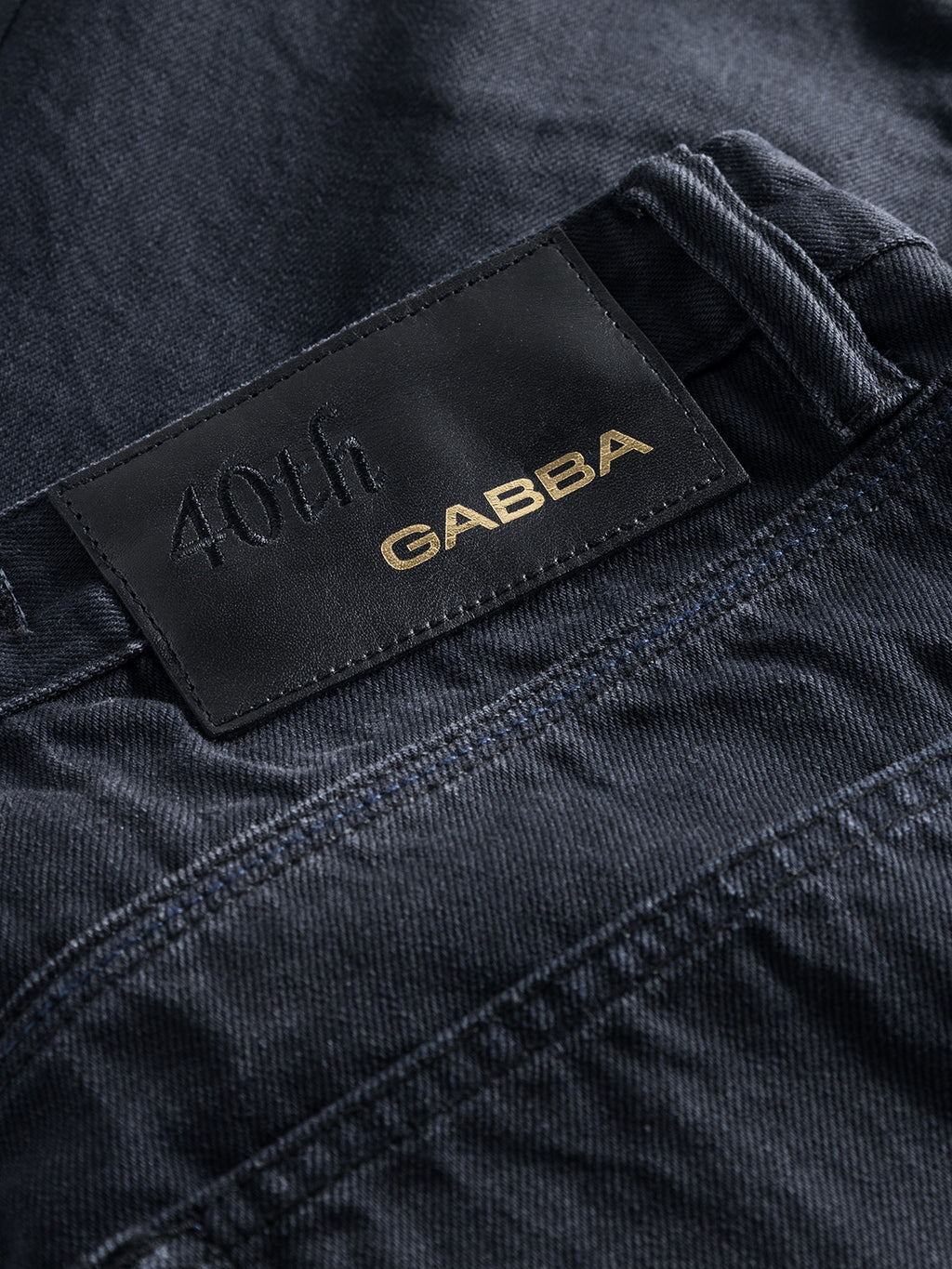 A close-up of the Carl Selvedge 40th Anniversary denim from Gabba shows a dark fabric with a black leather patch. The patch features 40th in black and GABBA in gold, emphasizing the unique marble appearance and durable quality of these special edition jeans.