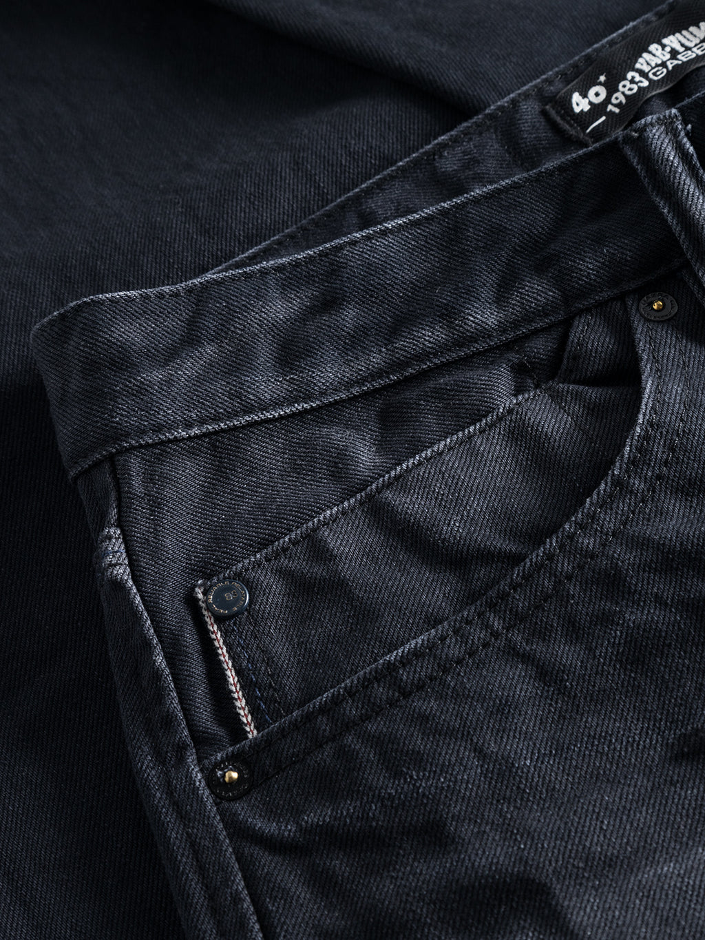 Close-up of Gabbas Carl Selvedge 40th Anniversary - Blue Black Denim jeans highlighting the waistband, front pocket, and partially visible button. The fabric shows a textured marble appearance, with part of a white label on the top right corner.