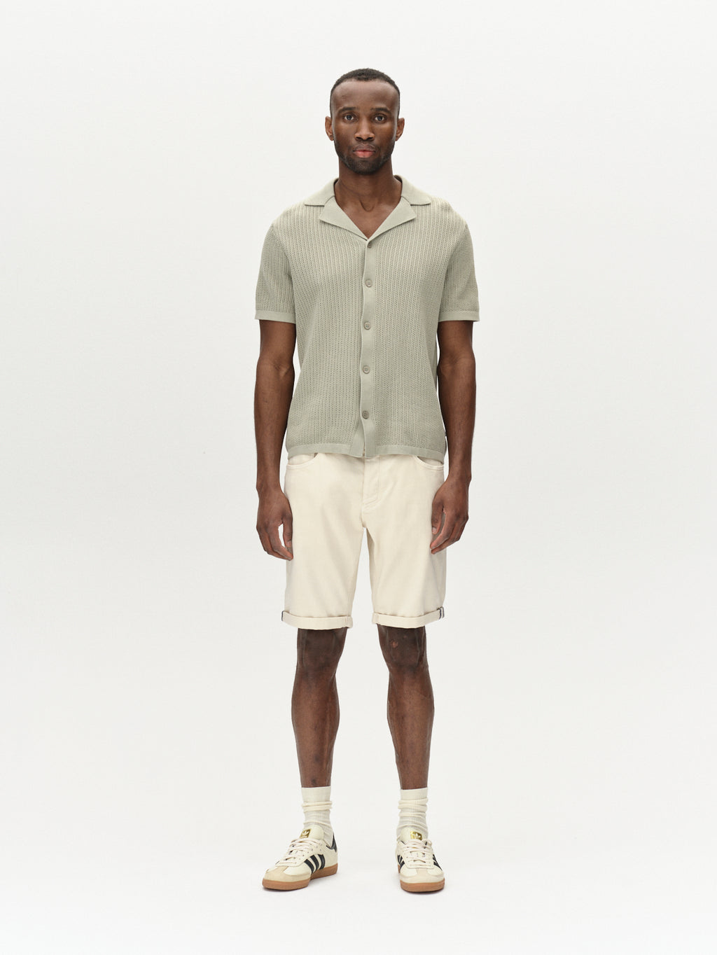 A person stands against a plain white background, wearing a light green short-sleeve button-up shirt and Gabbas Jason K3995 SANZA Shorts in Birch. Their look is completed with striped light-colored sneakers as they face the camera with their hands by their sides.