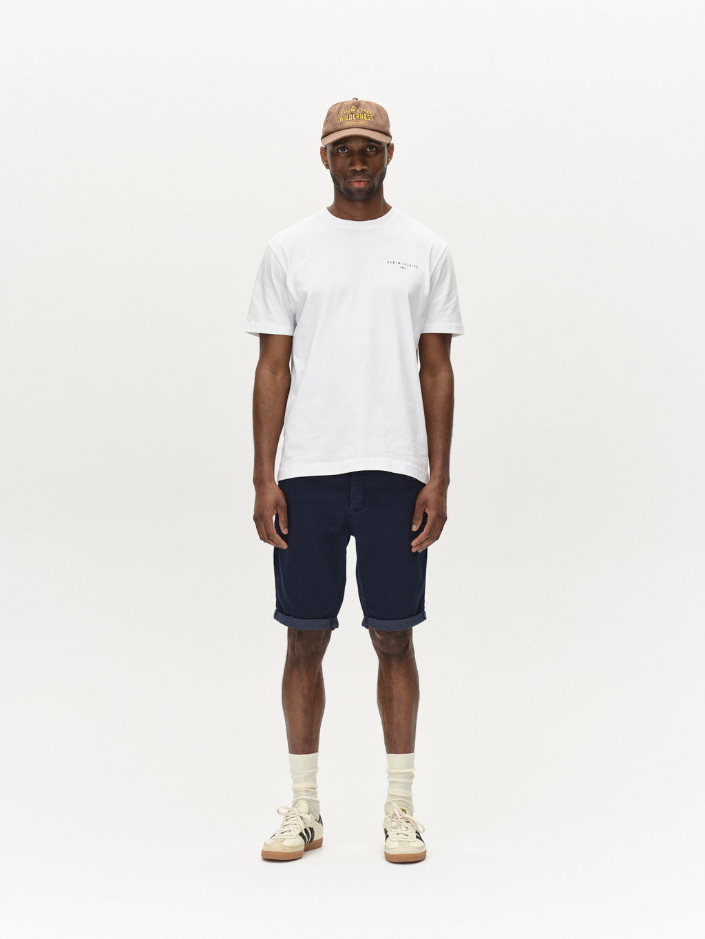 A person stands against a plain background, wearing a beige cap, white T-shirt, Gabbas Jason K3995 SANZA Shorts in navy, beige high-top sneakers, and white socks. They face the camera with a neutral expression and relaxed posture.