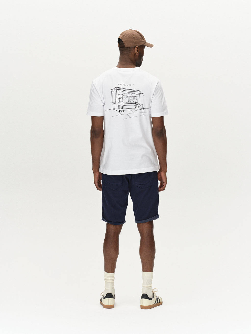 A person is wearing a beige cap, white graphic T-shirt with a line drawing on the back, and pairs perfectly with Gabbas Jason K3995 SANZA Shorts in navy. White socks and sneakers complete this versatile look against a plain background.