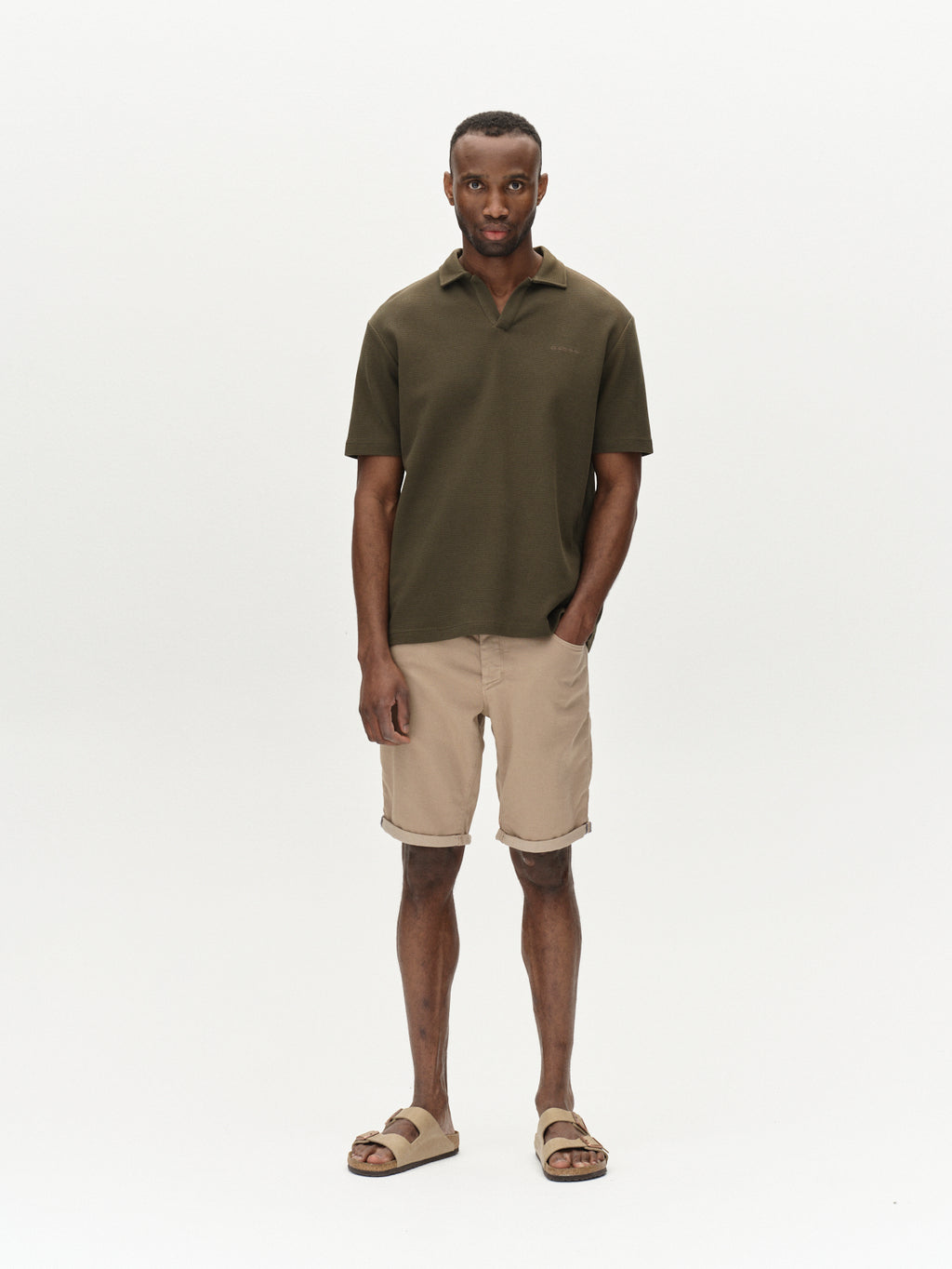 A man stands against a plain backdrop, wearing a dark green short-sleeve polo shirt and Gabbas Jason K3995 SANZA Shorts - Coriander. With one hand in his pocket, hes wearing beige sandals.