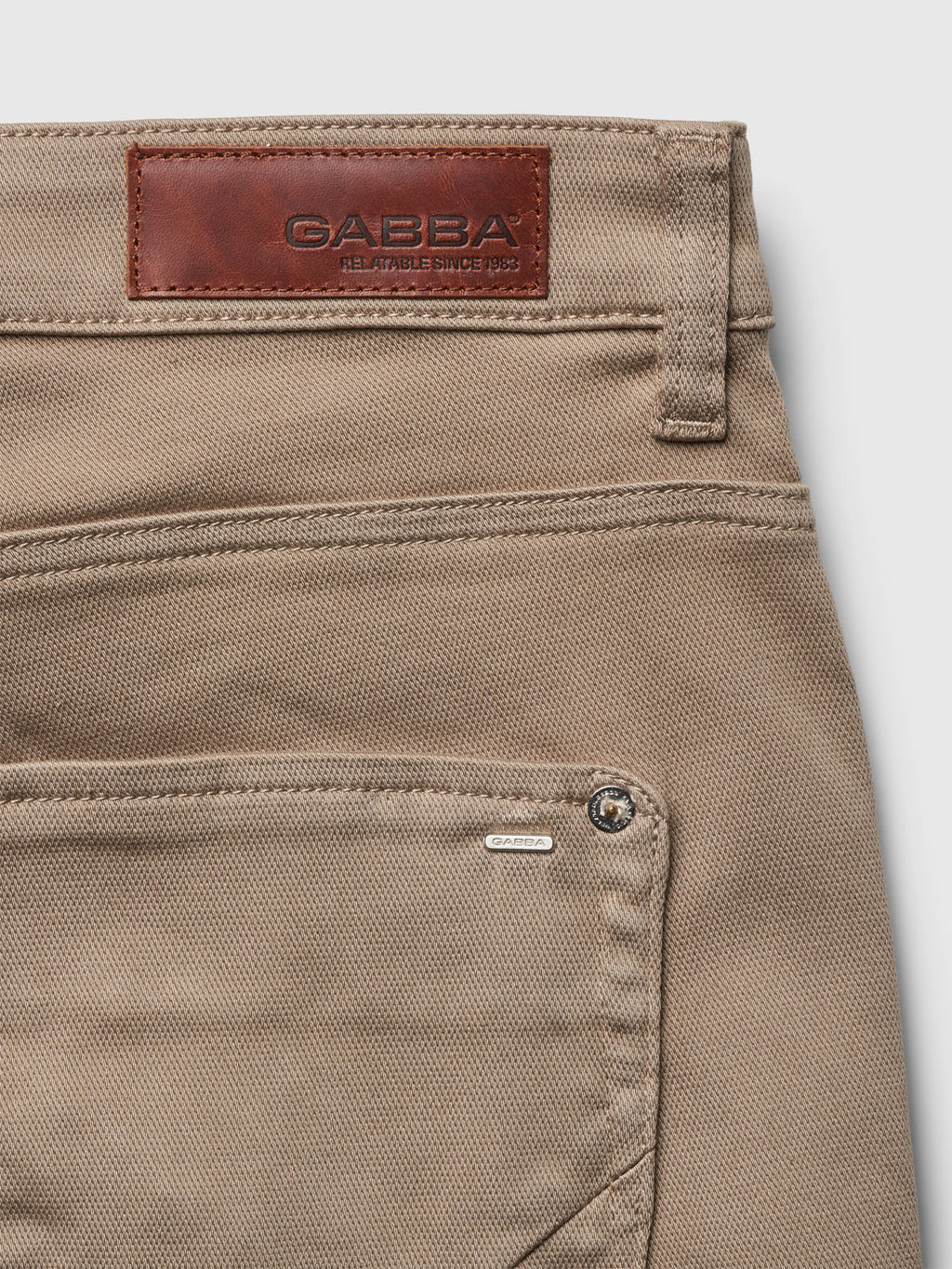 Close-up of beige denim fabric highlighting the top part of Gabbas Jason K3995 SANZA Shorts in Coriander, featuring a leather patch and metal tag on the front pocket. The super stretch fabric offers flexibility, making these shorts ideal for spring and summer adventures.