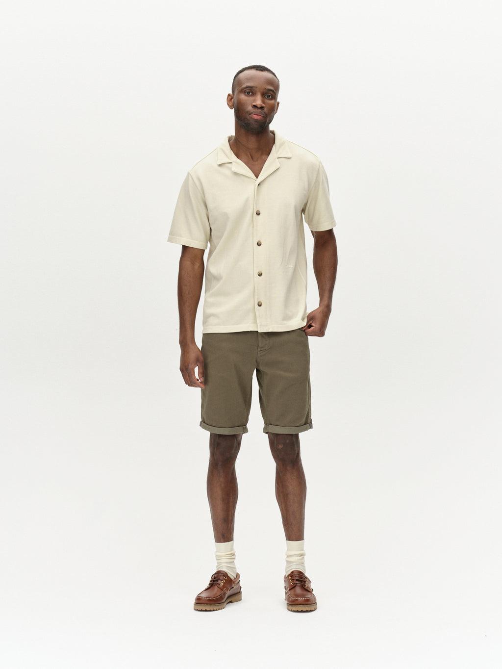 A man stands against a plain white background, wearing a beige short-sleeve button-up shirt, Gabbas Jason K3995 SANZA Shorts in Dusty Olive with reinforced pockets, beige socks, and brown shoes.
