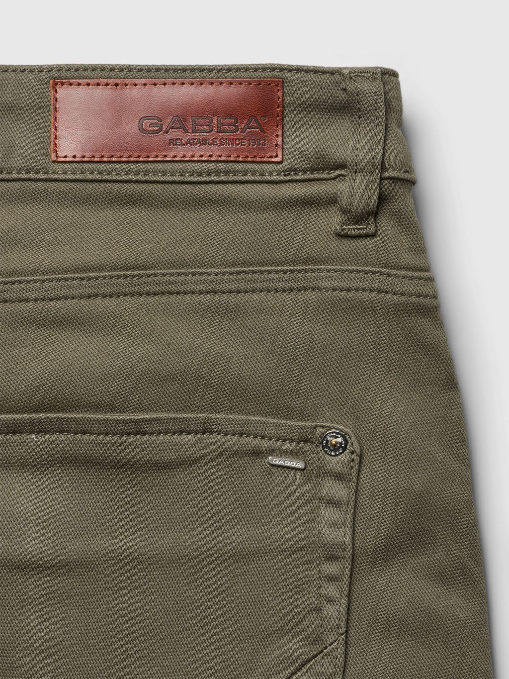 Check out the close-up of Jason K3995 SANZA Shorts in dusty olive by Gabba, made with super stretch fabric. They feature a brown leather patch with embossed text on the waistband, detailed stitching, and a visible front pocket with a small metallic button and tag.