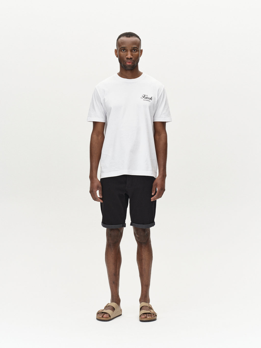 A person stands against a plain background wearing a white T-shirt with a small logo, Gabbas Jason K3995 SANZA knee-length denim shorts in black, and beige sandals. Their hands are at their sides as they face the camera, embodying effortless spring and summer style.