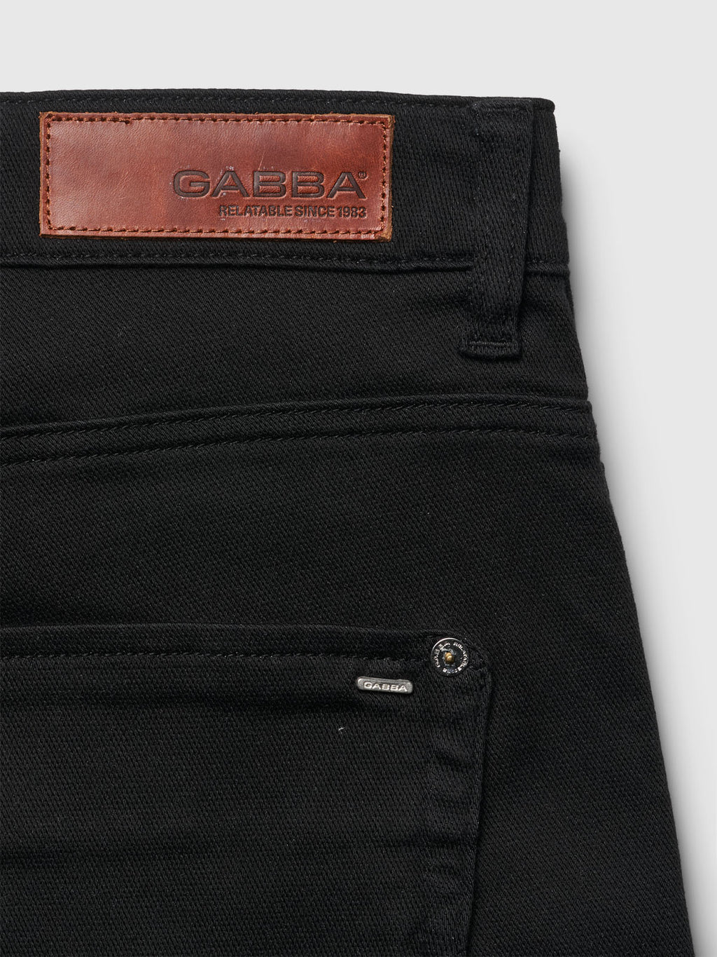 A close-up of Jason K3995 SANZA Shorts in black denim shows a brown leather label embossed with Gabba Reliable Since 1983 on the waistband. The shorts feature reinforced pockets, a small metal brand tag, super stretch fabric, and visible stitching details for enhanced comfort.