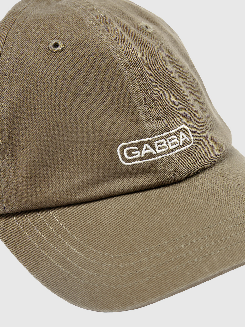 A close-up showcases the Gabba Cap - Rosin, a brown cap with GABBA embroidered in white. It features a classic design with a curved brim and metal eyelets for ventilation, ensuring comfort. The plain light gray background highlights its style.