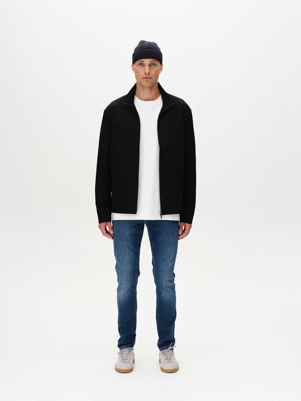 A man stands facing forward against a white background, wearing a black jacket, white t-shirt, Gabba Jones K3870 - Mid Blue Denim jeans with a slim fit, gray sneakers, and a dark knit beanie.
