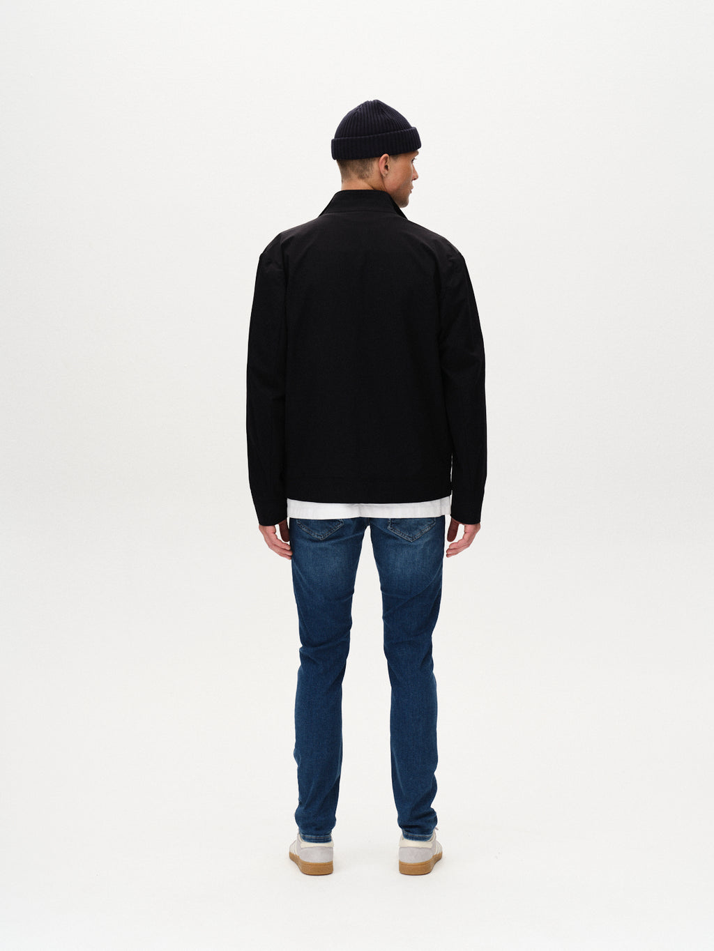 A man faces away from the camera, wearing Gabba Jones K3870 - Mid Blue Denim jeans with a slim silhouette, a black jacket, white sneakers, and a dark knit beanie against a plain white background.