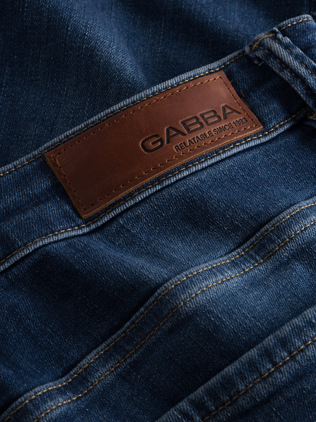 Close-up of folded Jones K3870 - Mid Blue Denim jeans with a brown leather brand label reading Gabba and Reliable Since 1983 on the waistband. The slim silhouette, reinforced pockets, and visible texture highlight the quality of the super-stretch fabric.