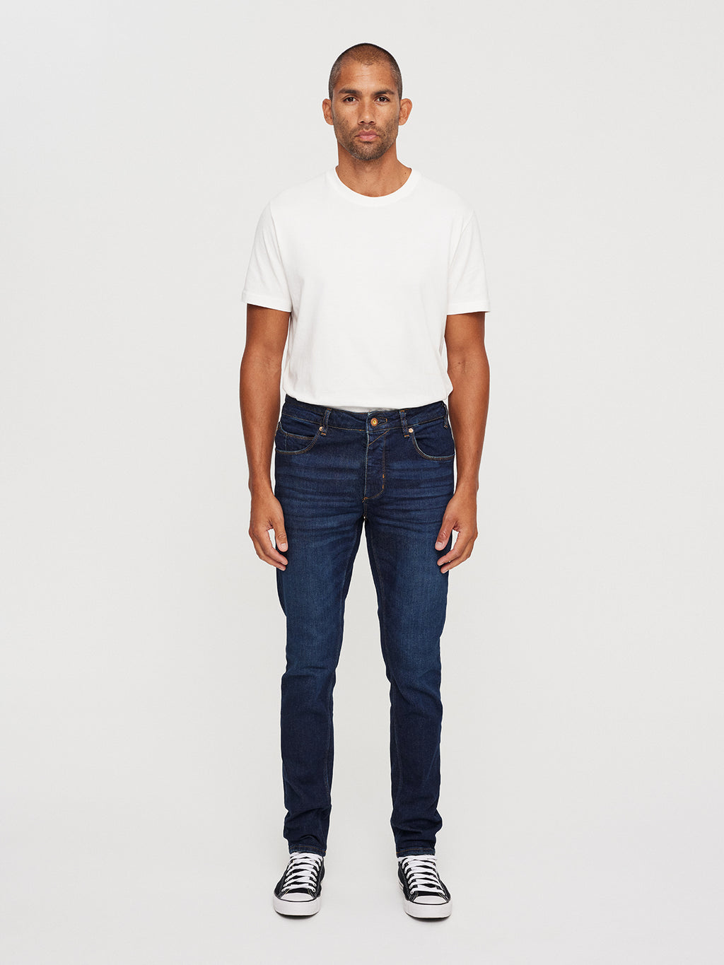A person with a slim appearance and laid-back look stands against a plain white background, wearing a white T-shirt, Rey K4441 Mid Blue Denim by Gabba, and black and white sneakers. They face forward with hands by their sides.