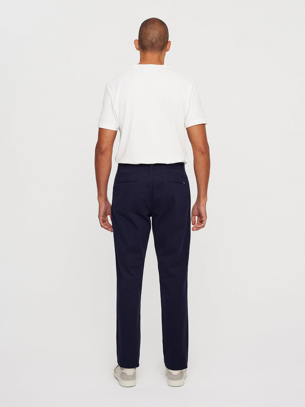 A person with a buzz cut faces away, wearing a classic white T-shirt, Gabbas Venza Chino in navy, and light-colored shoes against a white backdrop. This timeless look embodies the refined essentials of mens style.