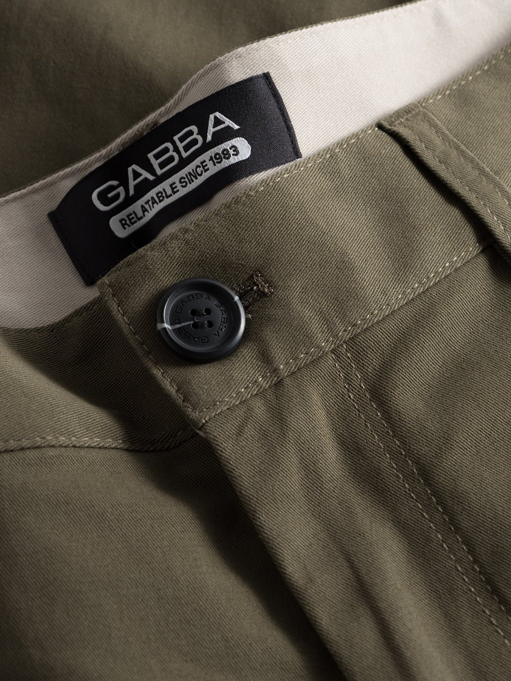 Close-up of the olive green Venza Chino pants with a black button and a black Gabba Reliable Since 1983 label sewn onto a light beige waistband, adding contrast to its earthy tone. Essential for any regular fit in mens wardrobes.