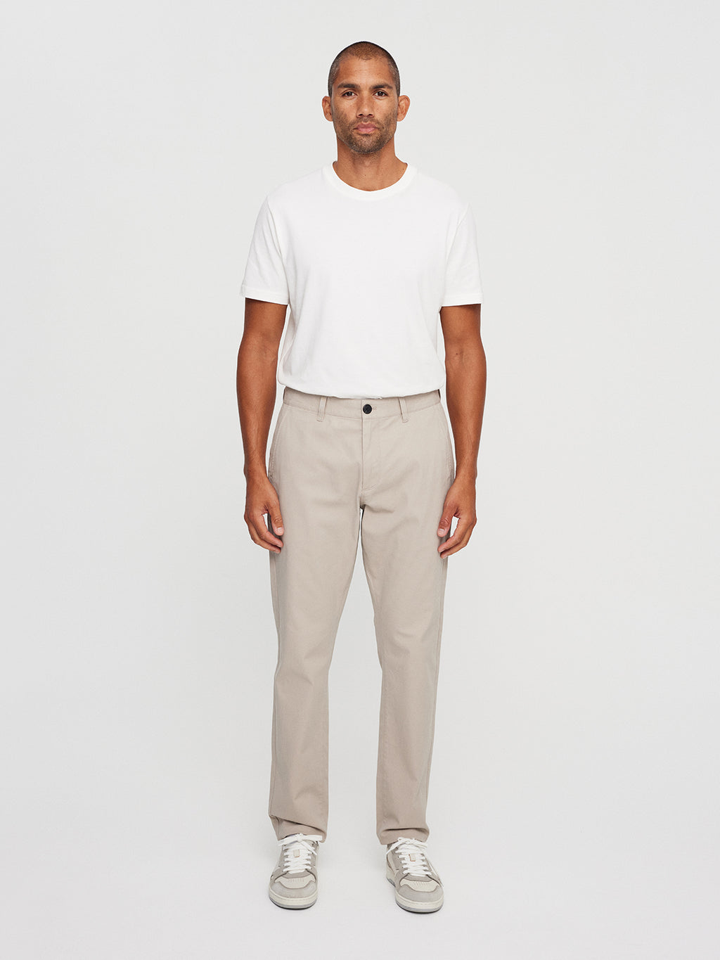 A person stands against a plain background wearing a white T-shirt, Venza Chino - Lt. Sand by Gabba, and white sneakers. With a neutral expression and relaxed arms, they embody a staple look for any mens wardrobe.