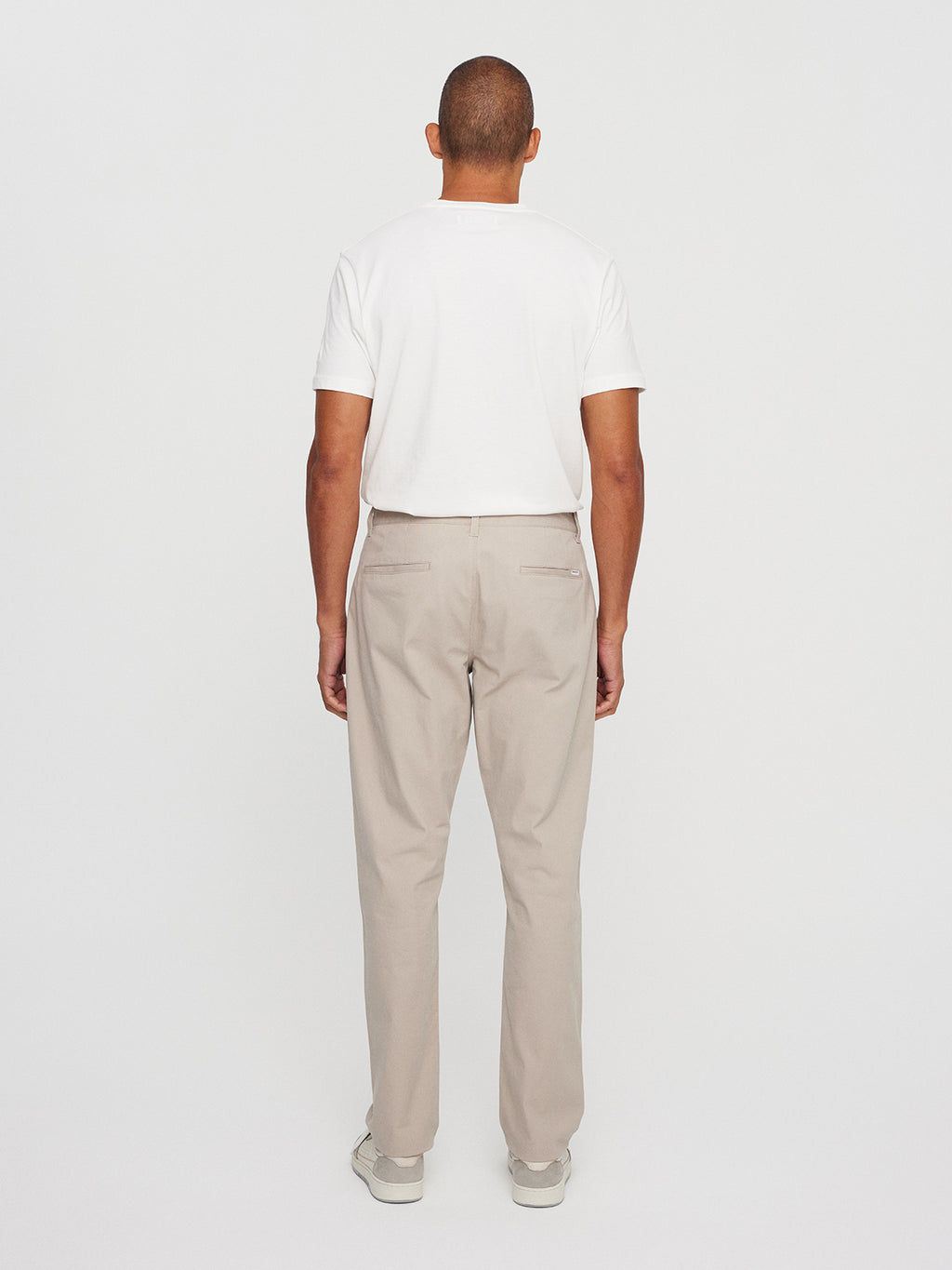 The individual, facing away from the camera, is modeling a crisp white T-shirt paired with Gabbas Venza Chino in Lt. Sand. Light-colored shoes complete their stylish ensemble against a plain white backdrop.