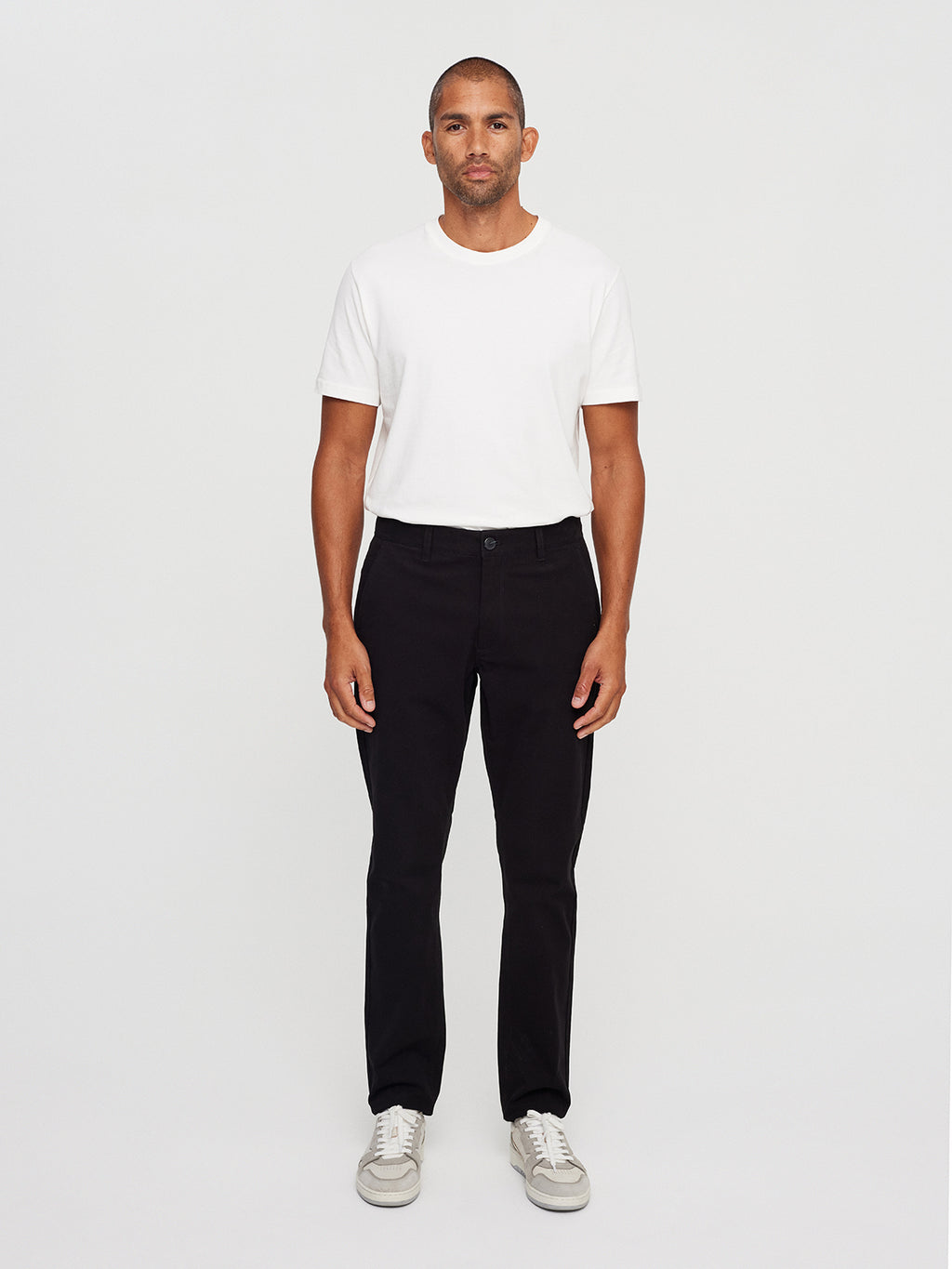 A person stands against a plain backdrop wearing a white T-shirt, Venza Chino - Black pants by Gabba, and white sneakers. They look directly at the camera with a neutral expression, highlighting an essential piece of mens wardrobe.