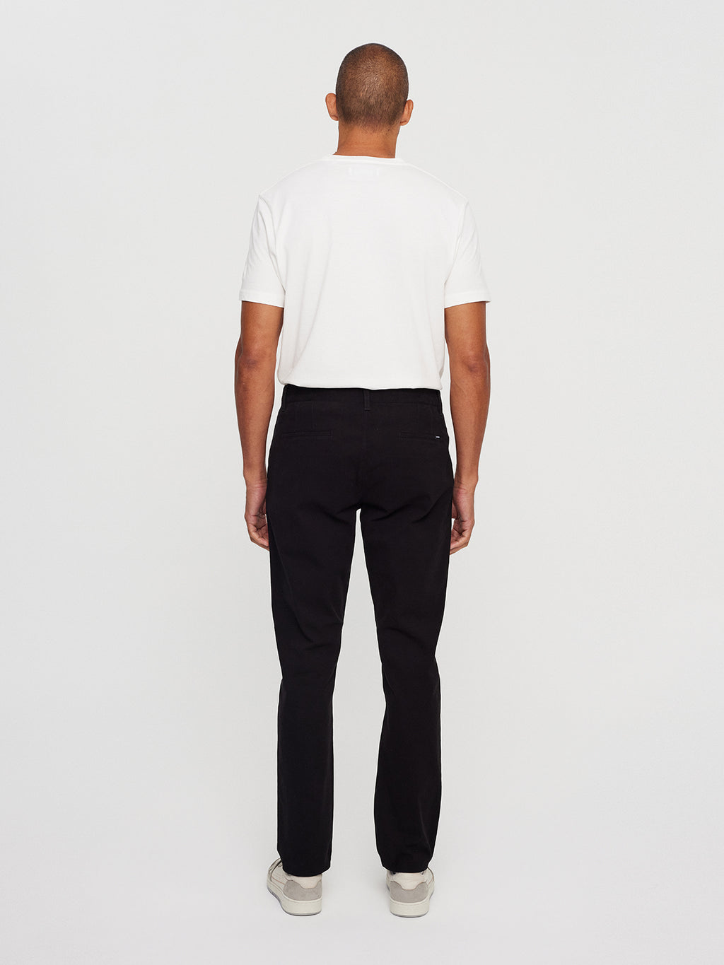 A person facing away, donning a plain white t-shirt and Venza Chino - Black by Gabba, paired with white shoes, stands against a plain white background.