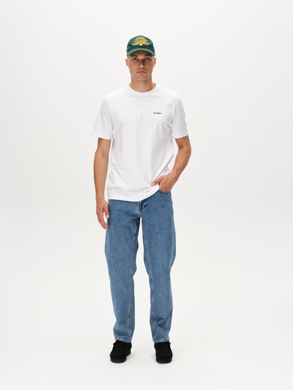 A man stands against a white background wearing the Gabba Dune Logo SS GOTS white t-shirt, paired with a green baseball cap, blue jeans, and black shoes. He has one hand in his pocket and a neutral expression.