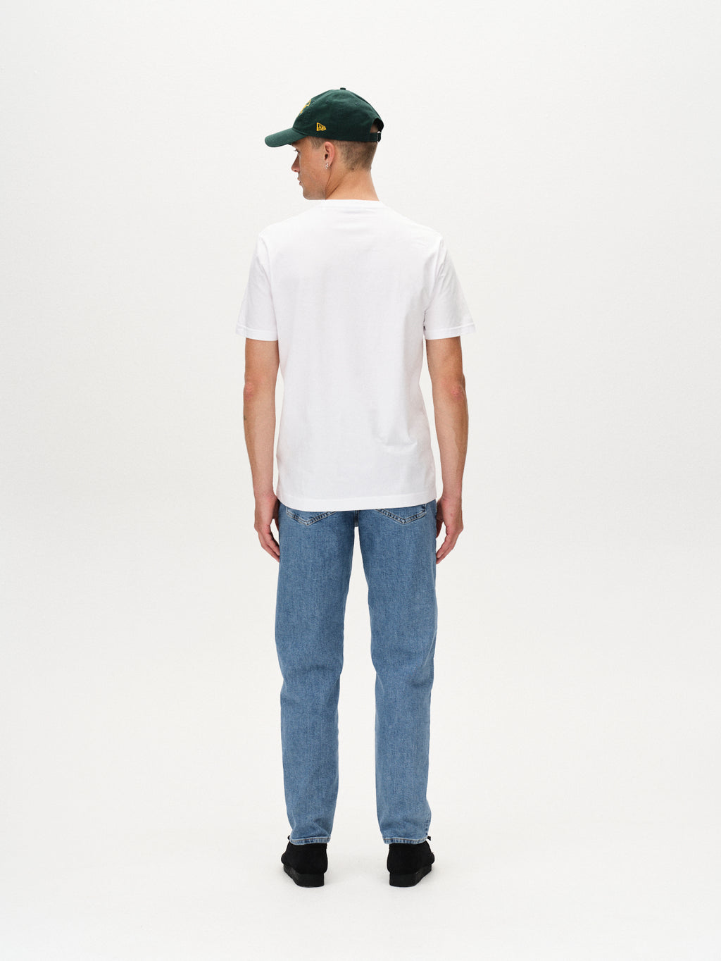 A person wearing the Gabba Dune Logo SS GOTS - White t-shirt, light blue jeans, black shoes, and a green cap stands facing away against a plain white background.