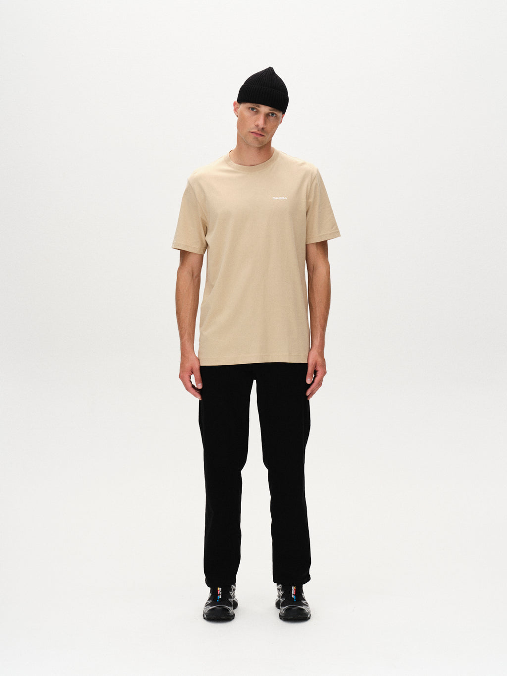A man stands against a plain white background, wearing the Gabba Dune Logo SS GOTS - Safari organic cotton beige t-shirt, paired with black pants, black beanie, and black shoes. His arms are relaxed at his sides and he has a neutral expression.