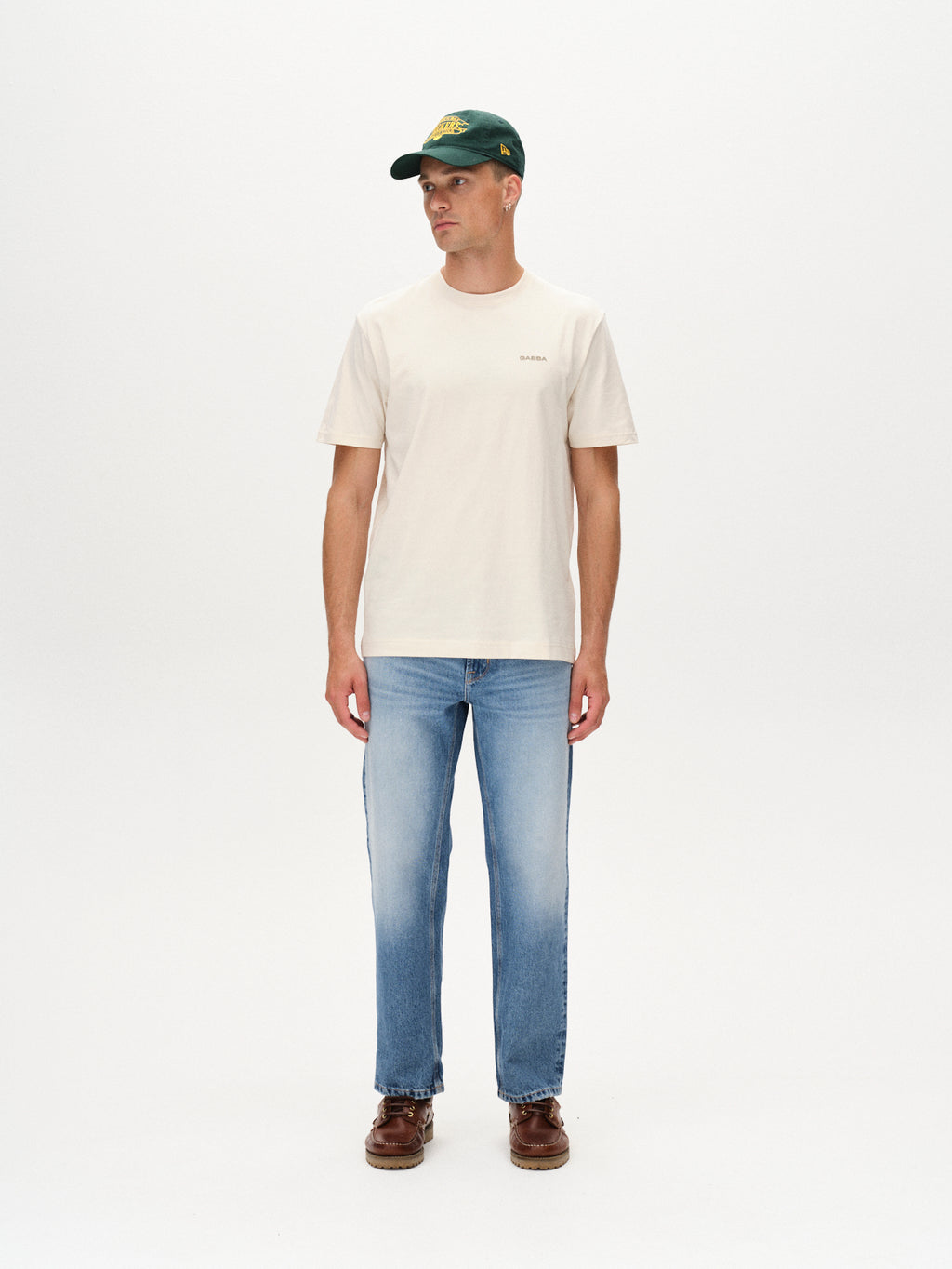 A man stands against a plain white background wearing the Gabba Dune Logo SS GOTS T-shirt in Birch, paired with blue jeans, brown shoes, and a dark green baseball cap, looking slightly to his left with his hands at his sides.