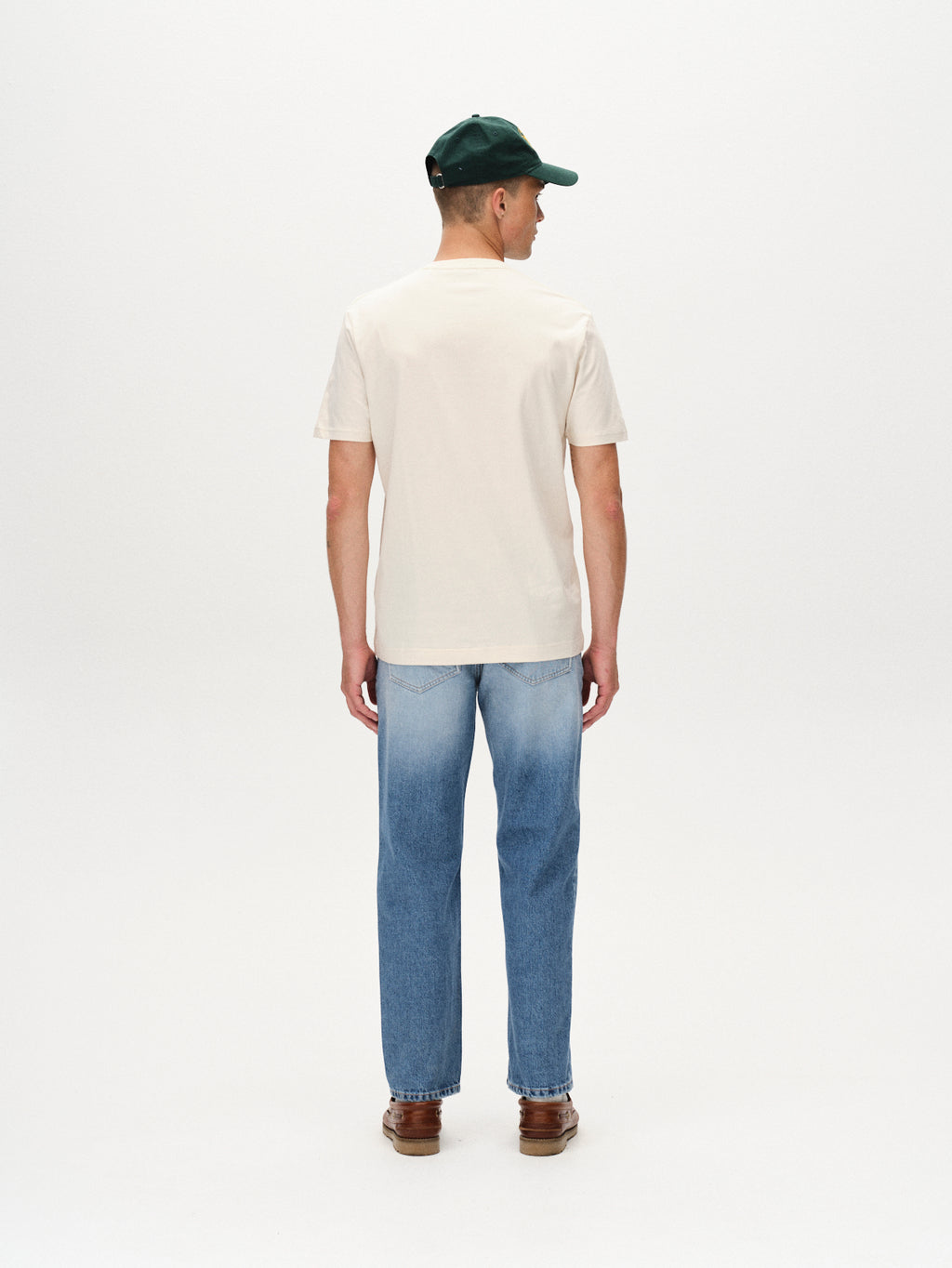A man stands facing away against a white background, wearing Gabbas Dune Logo SS GOTS - Birch cream organic cotton t-shirt, blue jeans, brown shoes, and a dark green cap.
