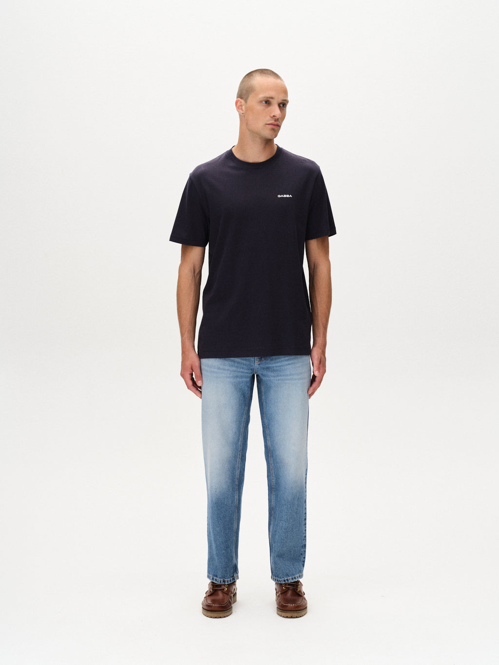 A man wears the Gabba Dune Logo SS GOTS t-shirt in Total Eclipse, paired with light blue jeans and brown shoes. He stands against a white background, looking slightly left with his arms relaxed at his sides.