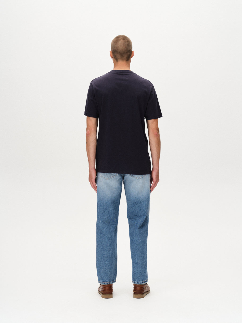 A person stands facing away from the camera, wearing the Gabba Dune Logo SS GOTS t-shirt in Total Eclipse, light blue jeans, and brown shoes against a plain white background.