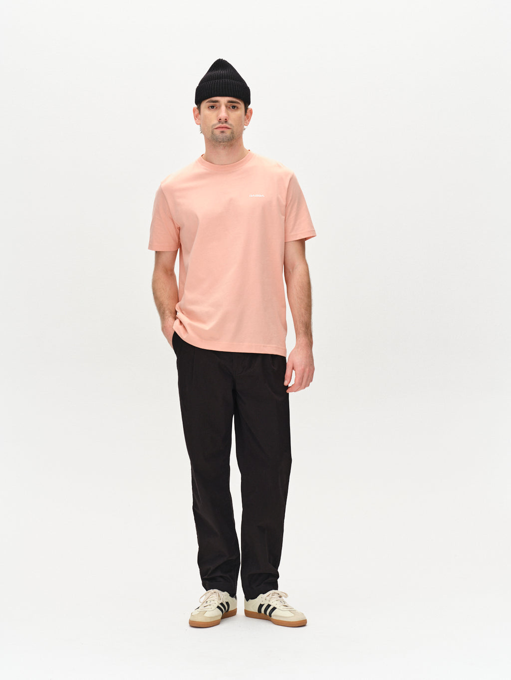 A person stands against a plain white background, wearing Gabbas Dune Logo SS GOTS - Rose Peach T-shirt, black pants, a black beanie, and white sneakers with black stripes.