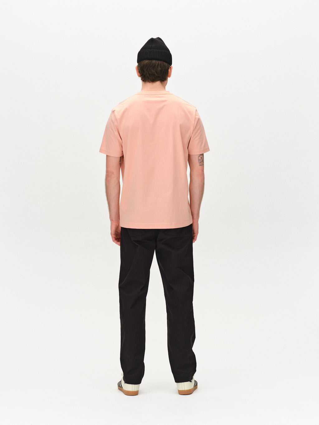 A person stands with their back to the camera against a plain white background, wearing a black beanie, Gabbas Dune Logo SS GOTS T-shirt in Rose Peach, black pants, and white shoes. Their arms hang relaxed by their sides.
