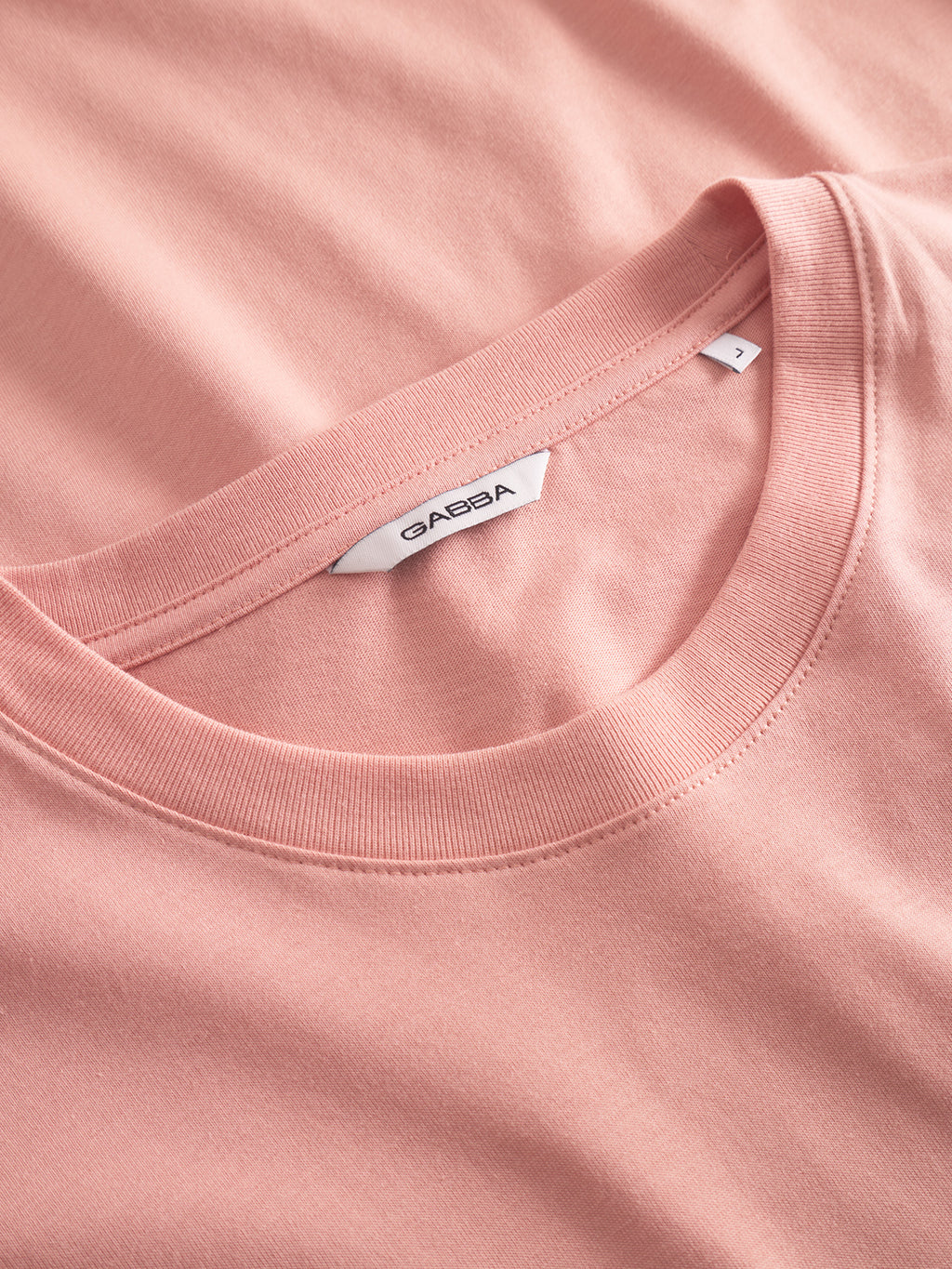 Close-up of a Dune Logo SS GOTS - Rose Peach T-shirt, made from organic cotton, featuring a white Gabba label near the neckline. The shirt is neatly laid on a similar pink fabric background, highlighting its texture and color.