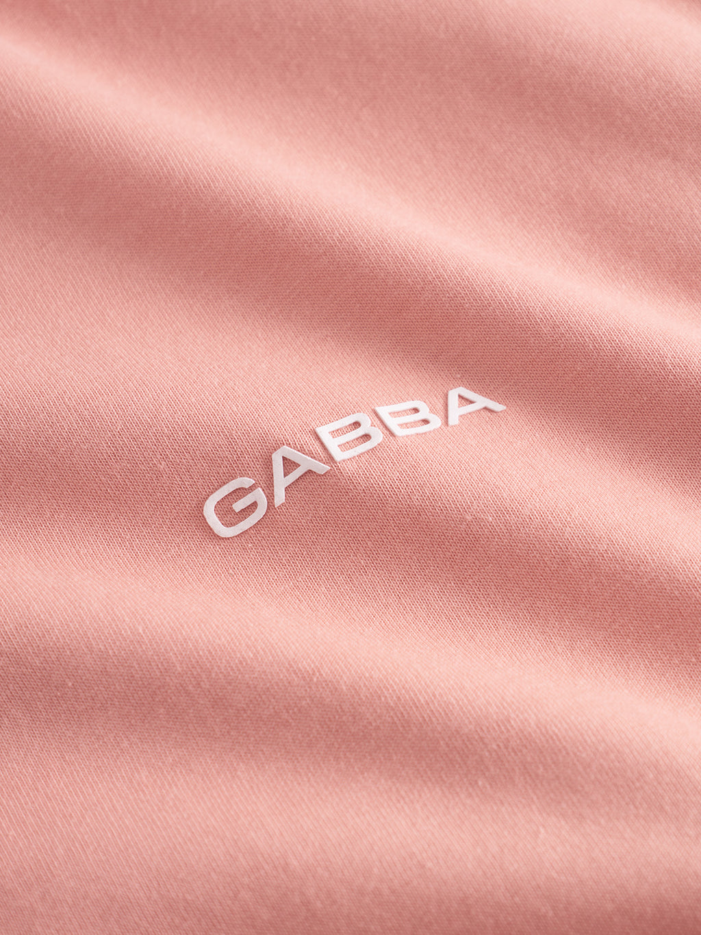 Close-up of the Dune Logo SS GOTS - Rose Peach by Gabba, made from organic cotton. The fabric showcases smooth waves and soft texture in light pink with GABBA in white letters, offering flexibility and an eco-friendly touch with its GOTS certification.